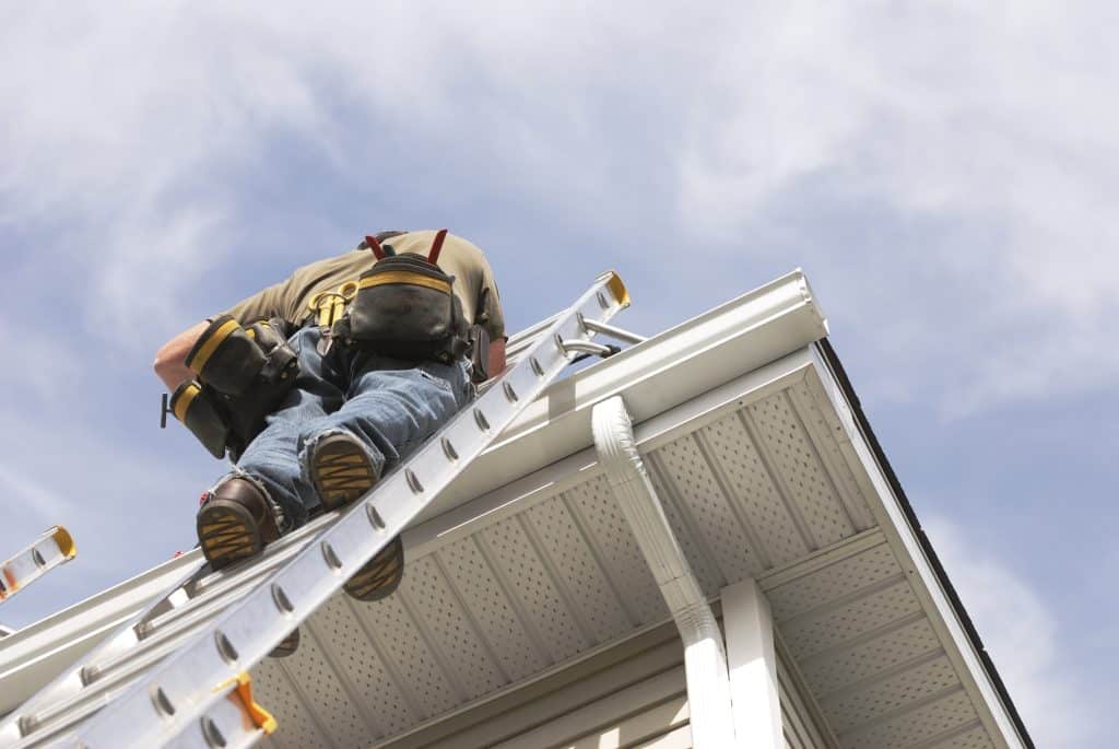 A handyman repairs his rain gutters. He is up a ladder, photo taken from ground looking up, low angle view. He wears a tool belt, sky and clouds, good copy space.