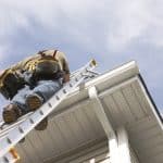 A handyman repairs his rain gutters. He is up a ladder, photo taken from ground looking up, low angle view. He wears a tool belt, sky and clouds, good copy space.